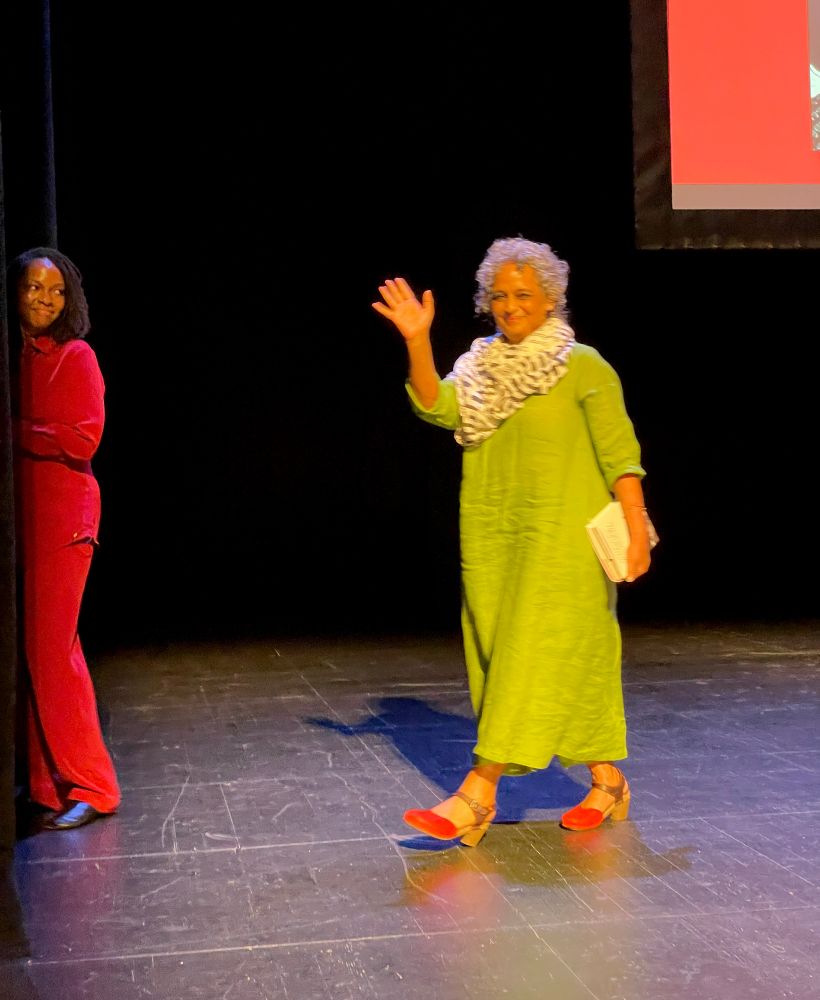 Arundahti Roy walks across the BAM stage on her way to the wings, waving to the audience. Imani Perry, already exiting, is looking back at her, smiling.