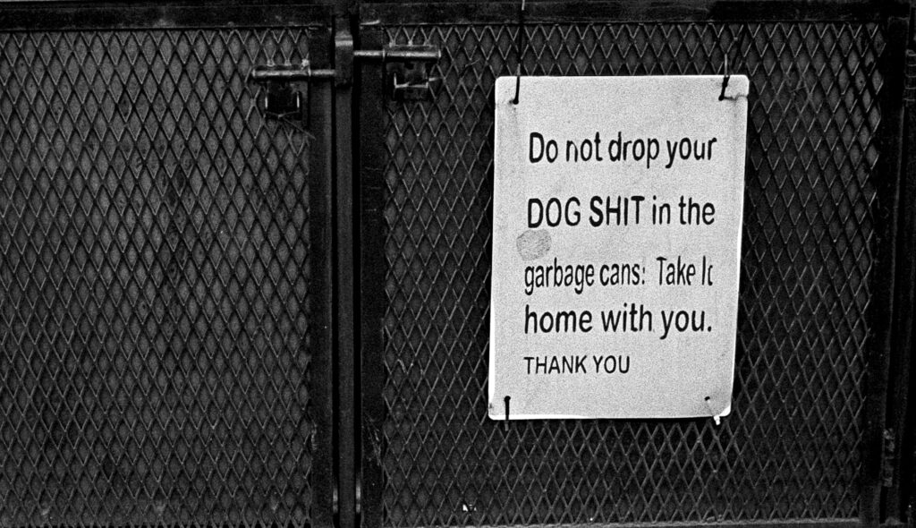 B&W photo of the side of a garbage tip with a sign reading "Do not drop your DOG SHIT in the garbage cans. Take it home with you. THANK YOU.