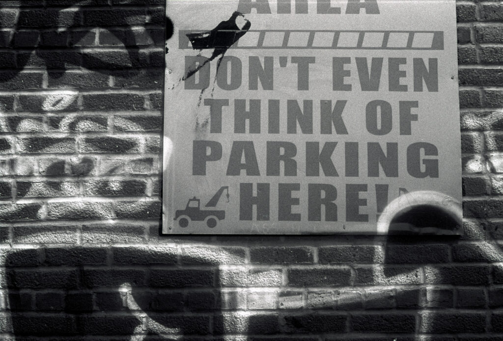 B&W 35mm photo of a graffiti covered wall with a sign reading "DON'T EVEN THINK OF PARKING HERE"