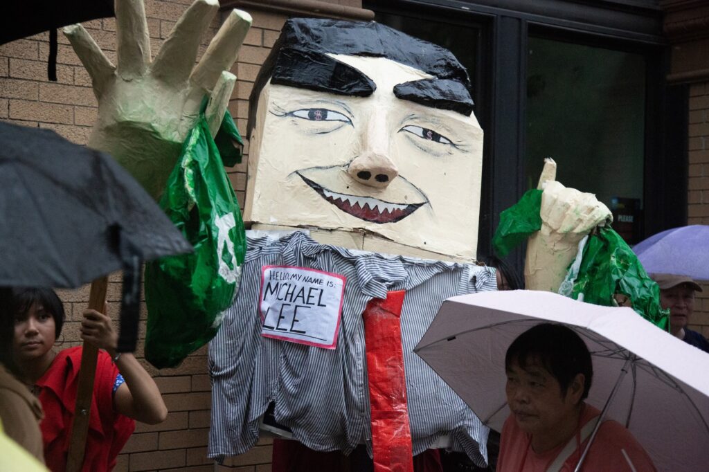 Protestors with umbrellas in front of a giant, larger than life handmade puppet of a figure with jagged teeth and holding what appear to be bags of money. Nametag on the puppet reads "Hello, my name is Michael Lee"