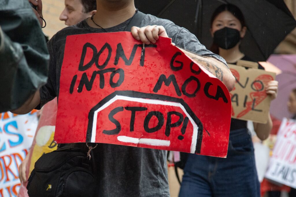 Protestor in the rain holds up a red hand made sign reading "DON'T GO INTO MOCA / STOP!"