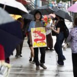 Protestors from Youth Against Displacement march in the rain holding umbrellas outside of the Museum of Chinese in America town hall. A protestor's sign reads "BOYCOTT MOCA" and features a red dragon drawing.