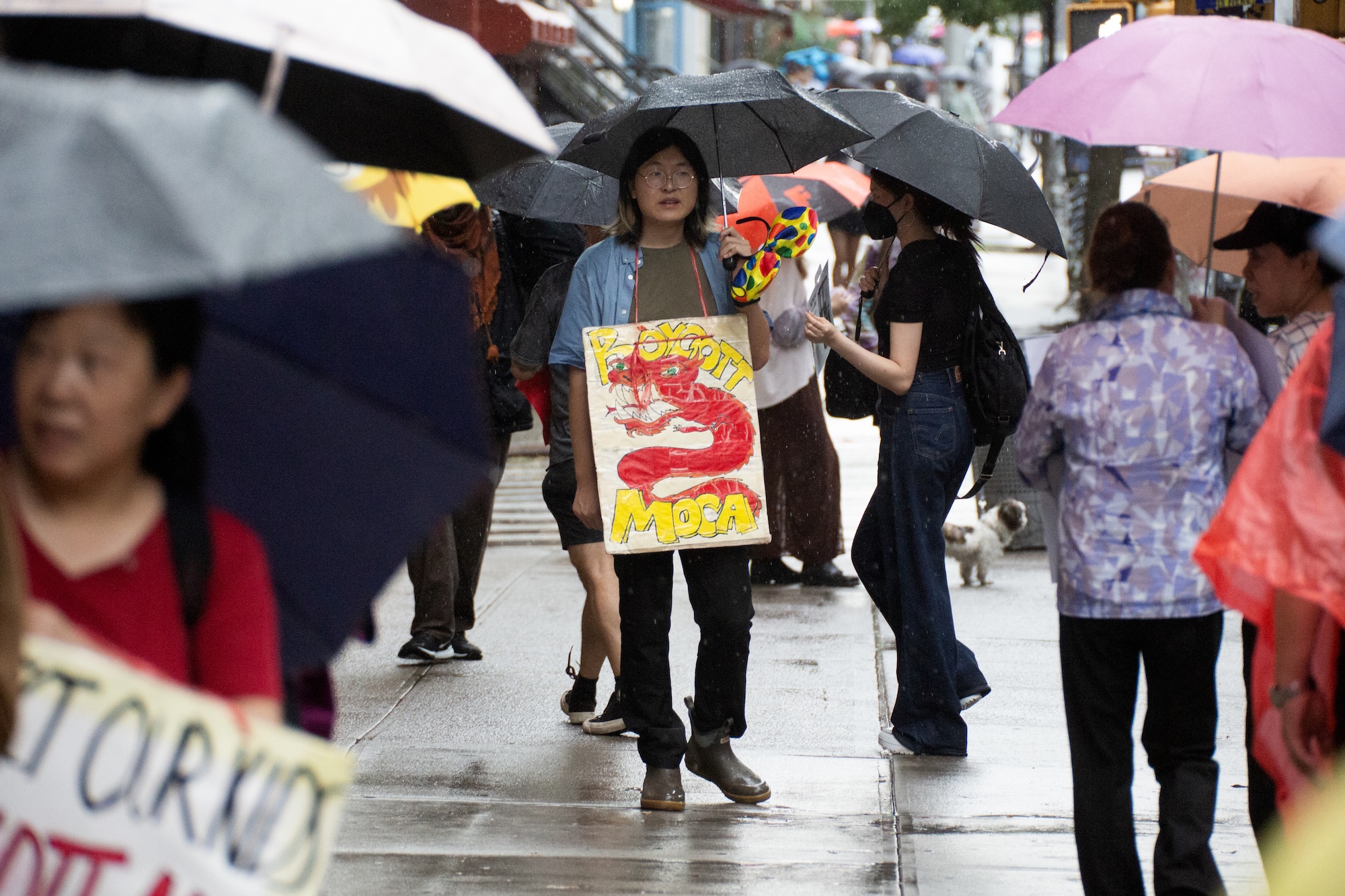 Protestors from Youth Against Displacement march in the rain holding umbrellas outside of the Museum of Chinese in America town hall. A protestor's sign reads "BOYCOTT MOCA" and features a red dragon drawing.
