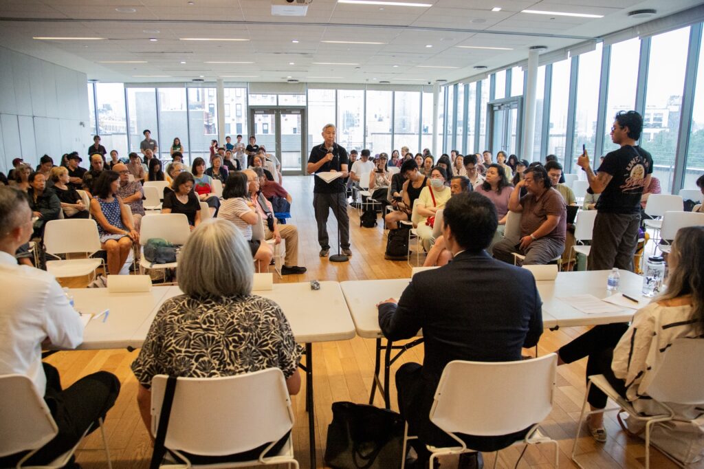 Wide shot of the MOCA town hall, seen from behind the panelists. Rocky Chin stands in the aisle between the chairs of the audience, speaking into a mic