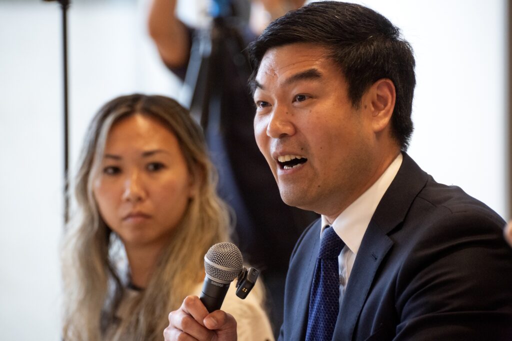 MOCA president Michael Lee speaks into a microphone at the town hall as Vic Lee watches him.