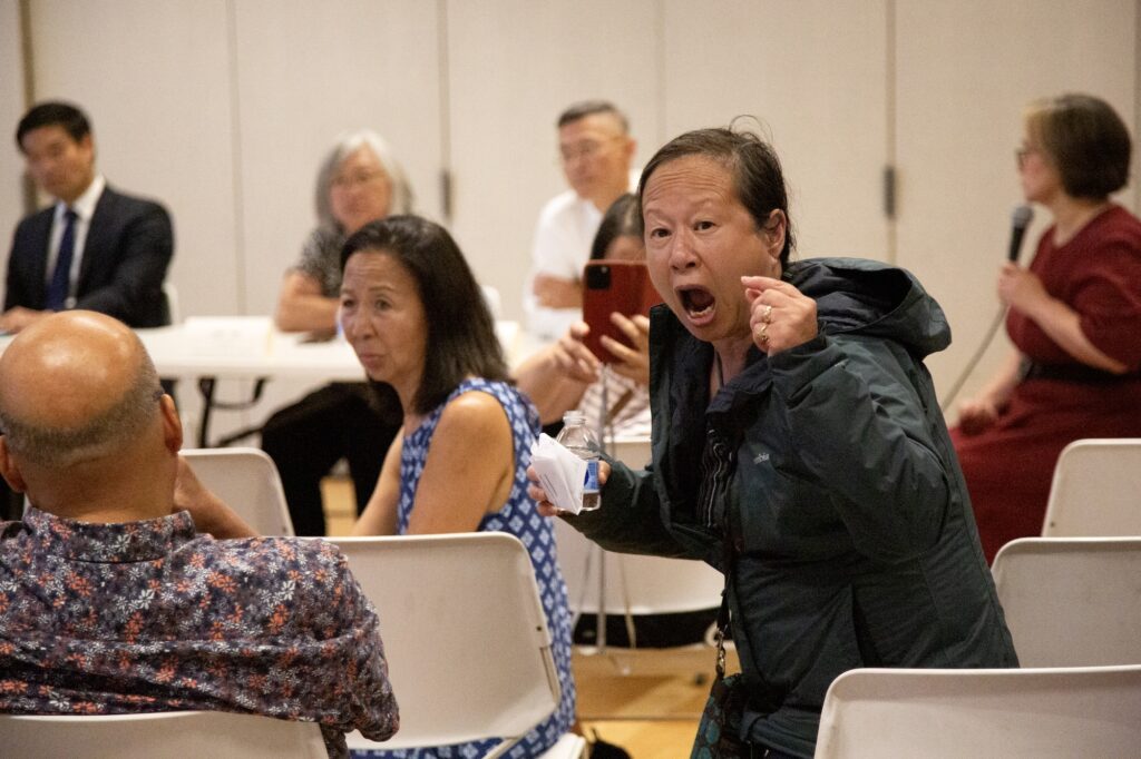 Liz Ouyang stands at the town hall, turning and shouting, one hand raised