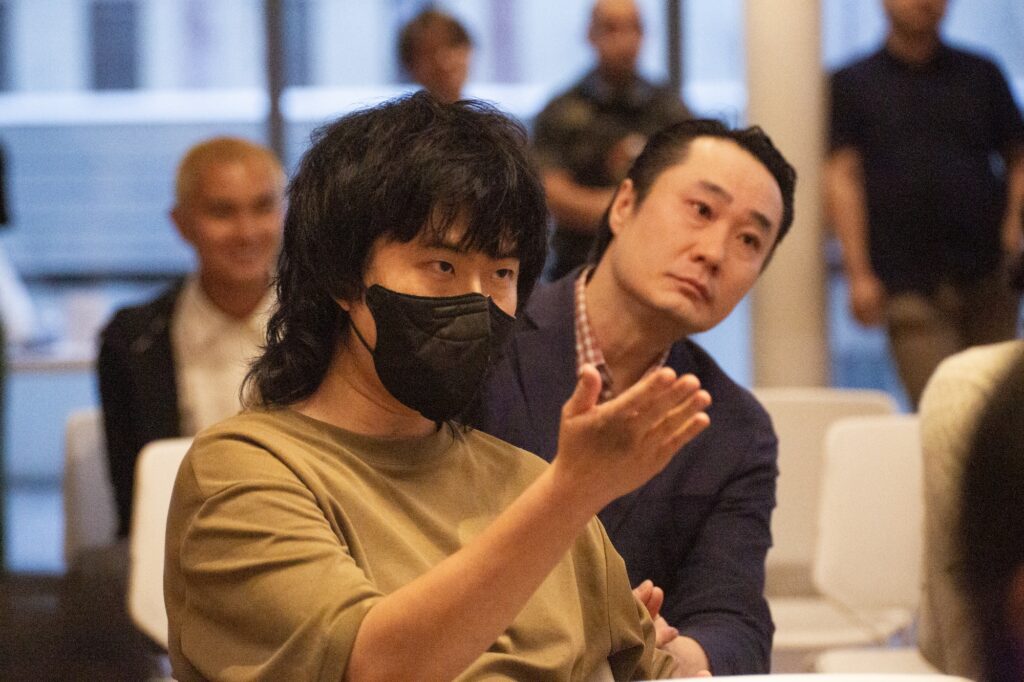 Aaron Yin sits at the town hall, wearing a mask, one hand raised as he responds