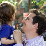 David Siffert holds their toddler, beaming at them. The toddler has their face turned away from us, looking at something in the background.