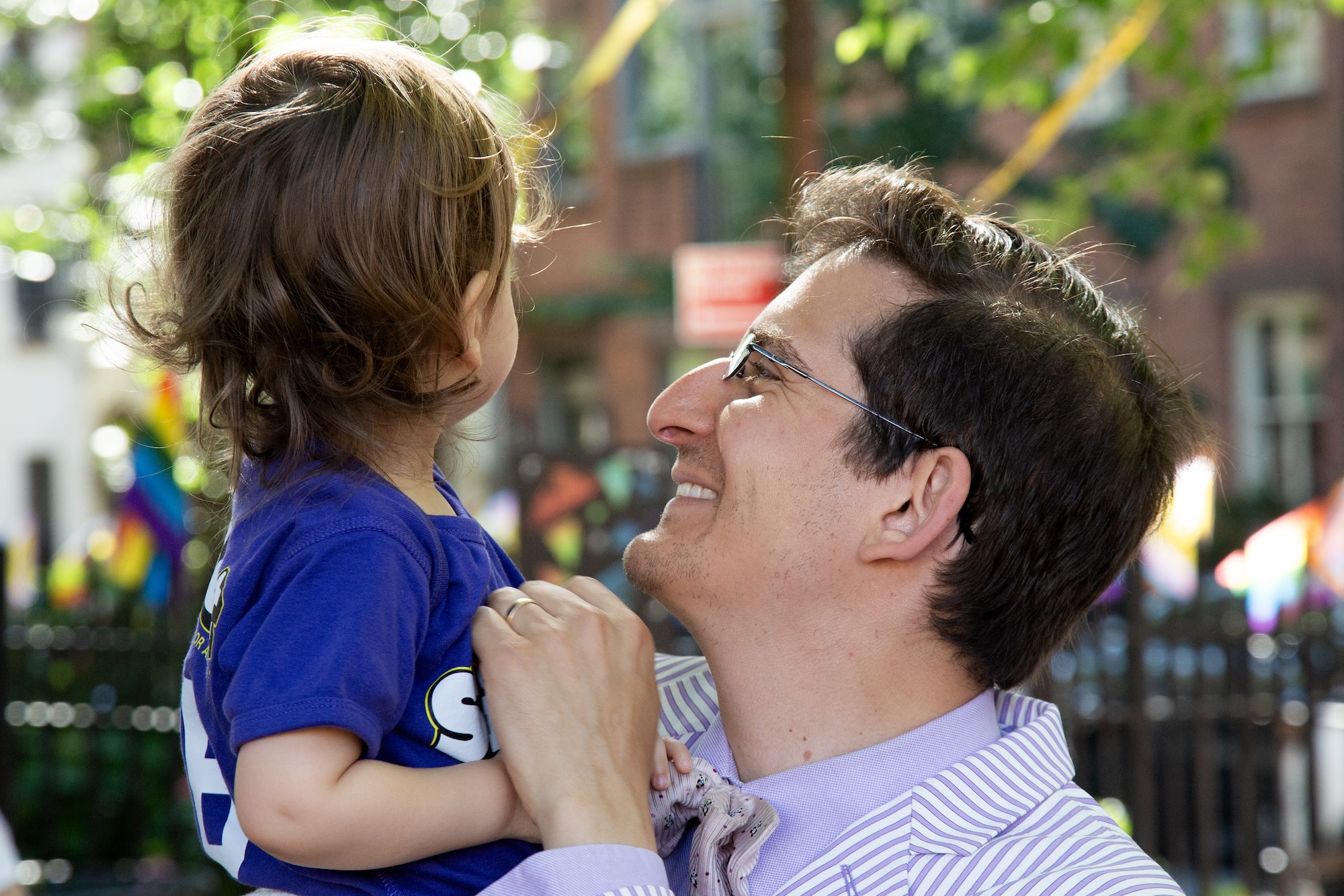 David Siffert holds their toddler, beaming at them. The toddler has their face turned away from us, looking at something in the background.