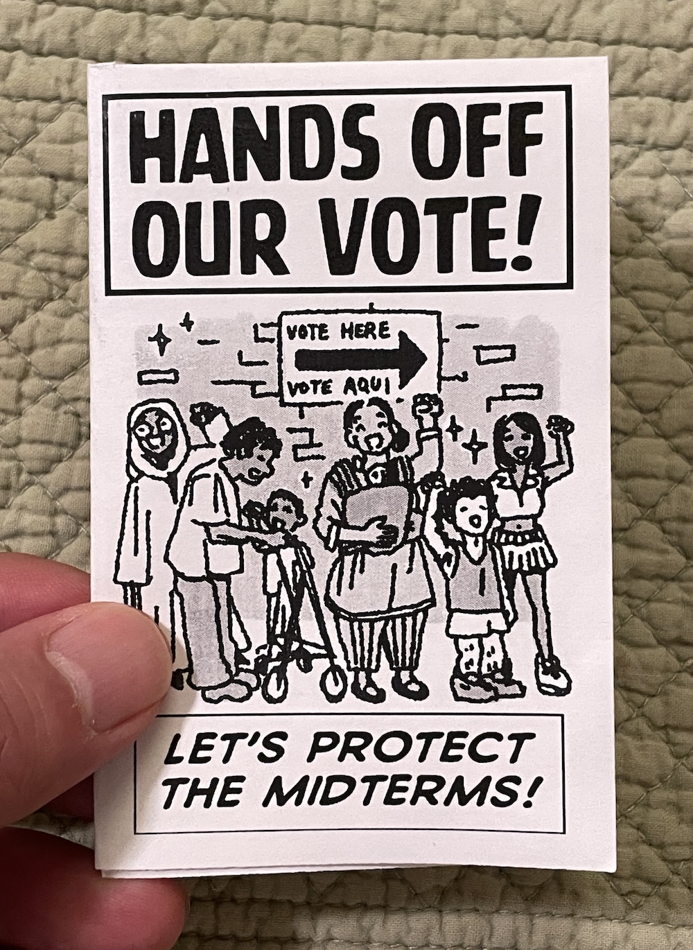Hand holding a zine titled "HANDS OFF OUR VOTE!" with the subtitle "LET'S PROTECT THE MIDTERMS!" Art by Priya Huq shows a multiracial group of people of various ages standing lined up in front of a "VOTE HERE / VOTE AQUI" sign, smiling, with fists raised.